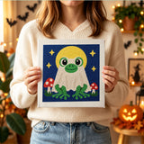Tableau punch needle grenouille lune tenu en main pour montrer sa taille